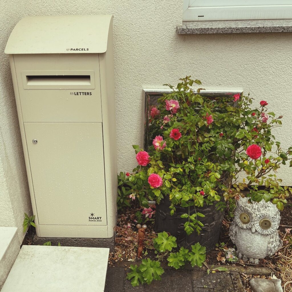 Extra large cream slanted top smart parcel box next to a flower bush and an ornamental owl.